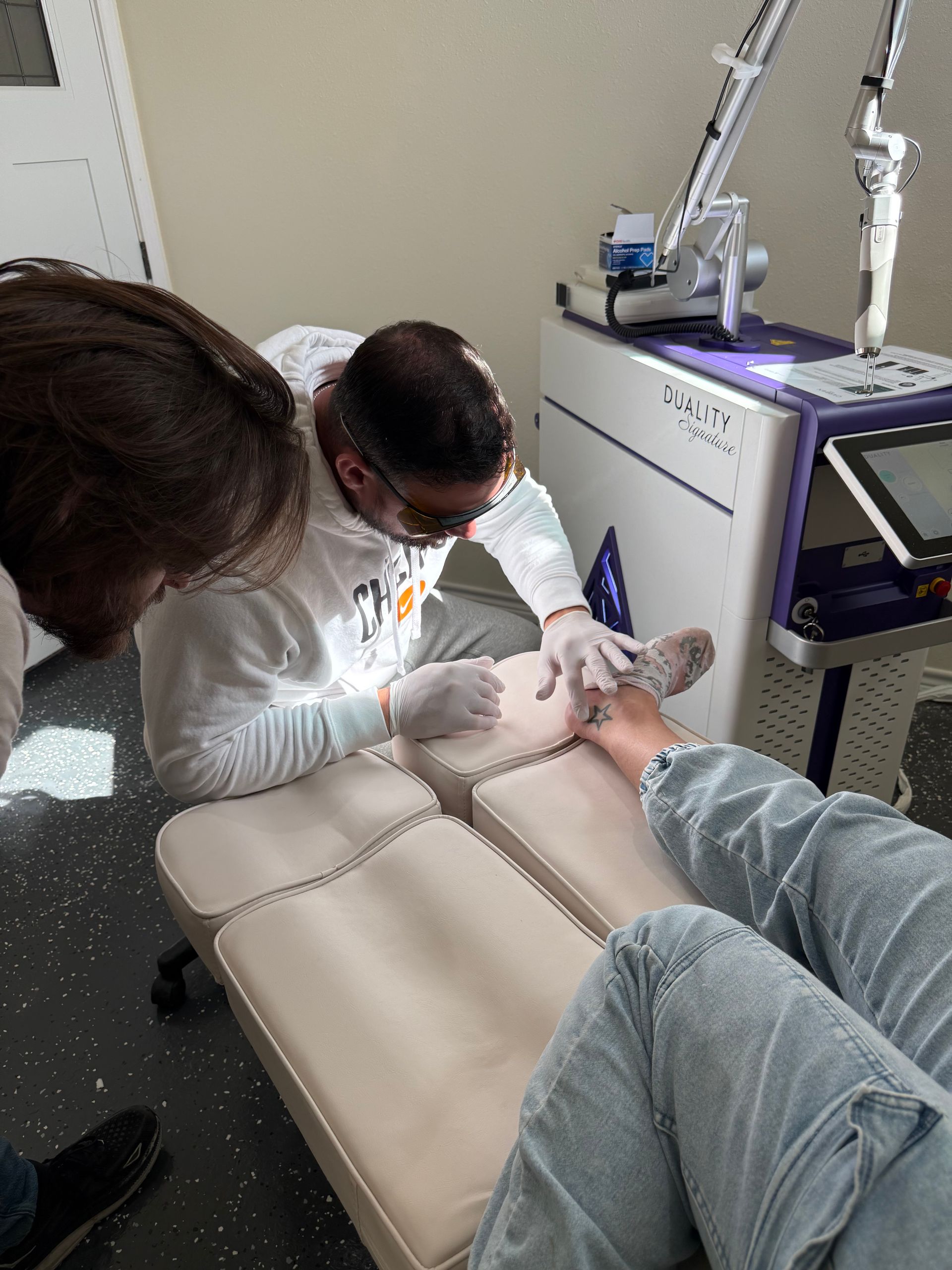 Person using laser tattoo removal device on a patient's leg in a clinic. Purple and white equipment.