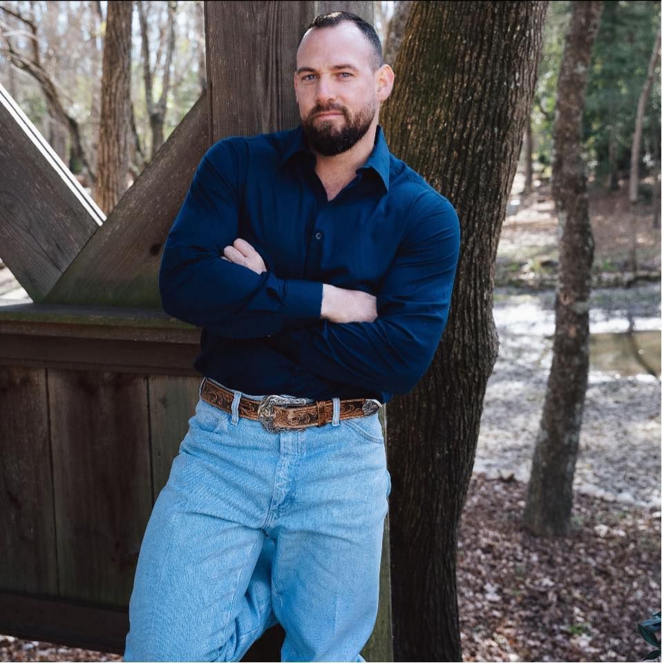 Man with beard, arms crossed, leaning against wooden structure and tree, wearing blue shirt and jeans. Outdoors.