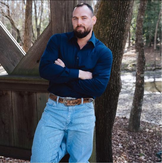 Man with beard, arms crossed, leaning against wooden structure and tree, wearing blue shirt and jeans. Outdoors.