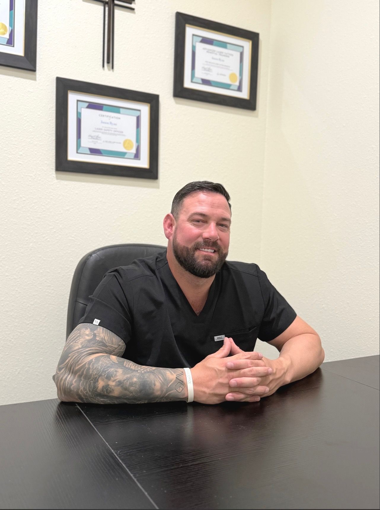 Man with beard, arms crossed, leaning against wooden structure and tree, wearing blue shirt and jeans. Outdoors.