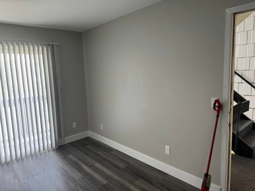 Empty room with gray walls, wooden floors, vertical blinds, and a doorway leading to stairs.