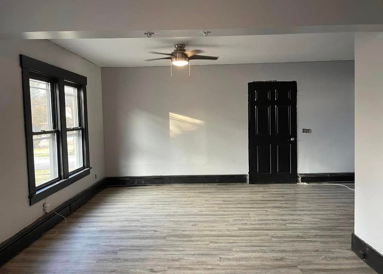 Empty living room with gray walls, dark trim, and black door. Window with black frame on the left. Gray wood floor.