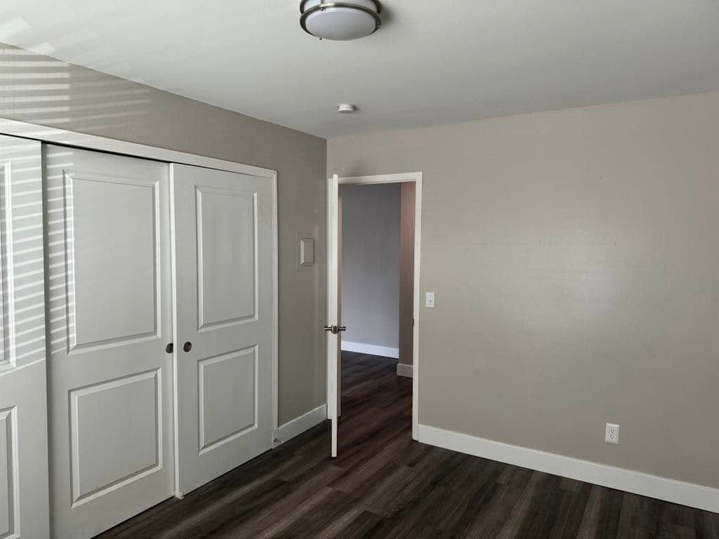 Bedroom with white closet doors, gray walls, and dark wood-look flooring. An open doorway leads to another room.