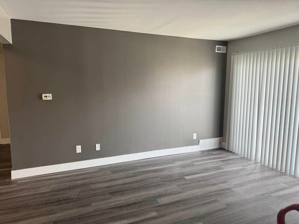 Empty living room with gray walls, white trim, gray wood-look floors, and vertical blinds.