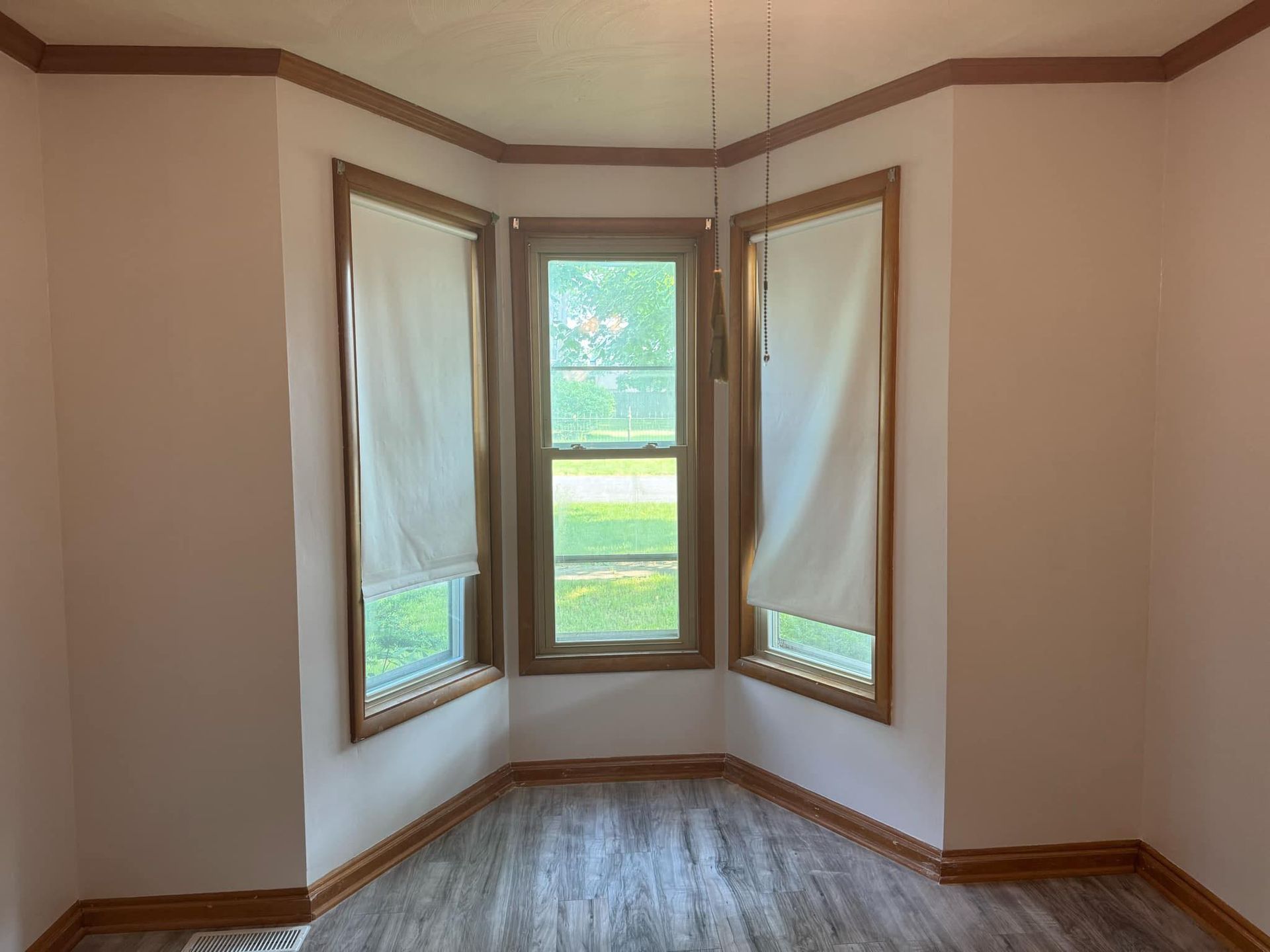 Bay window with beige walls, white shades, and wood trim in a room with wood-look flooring.