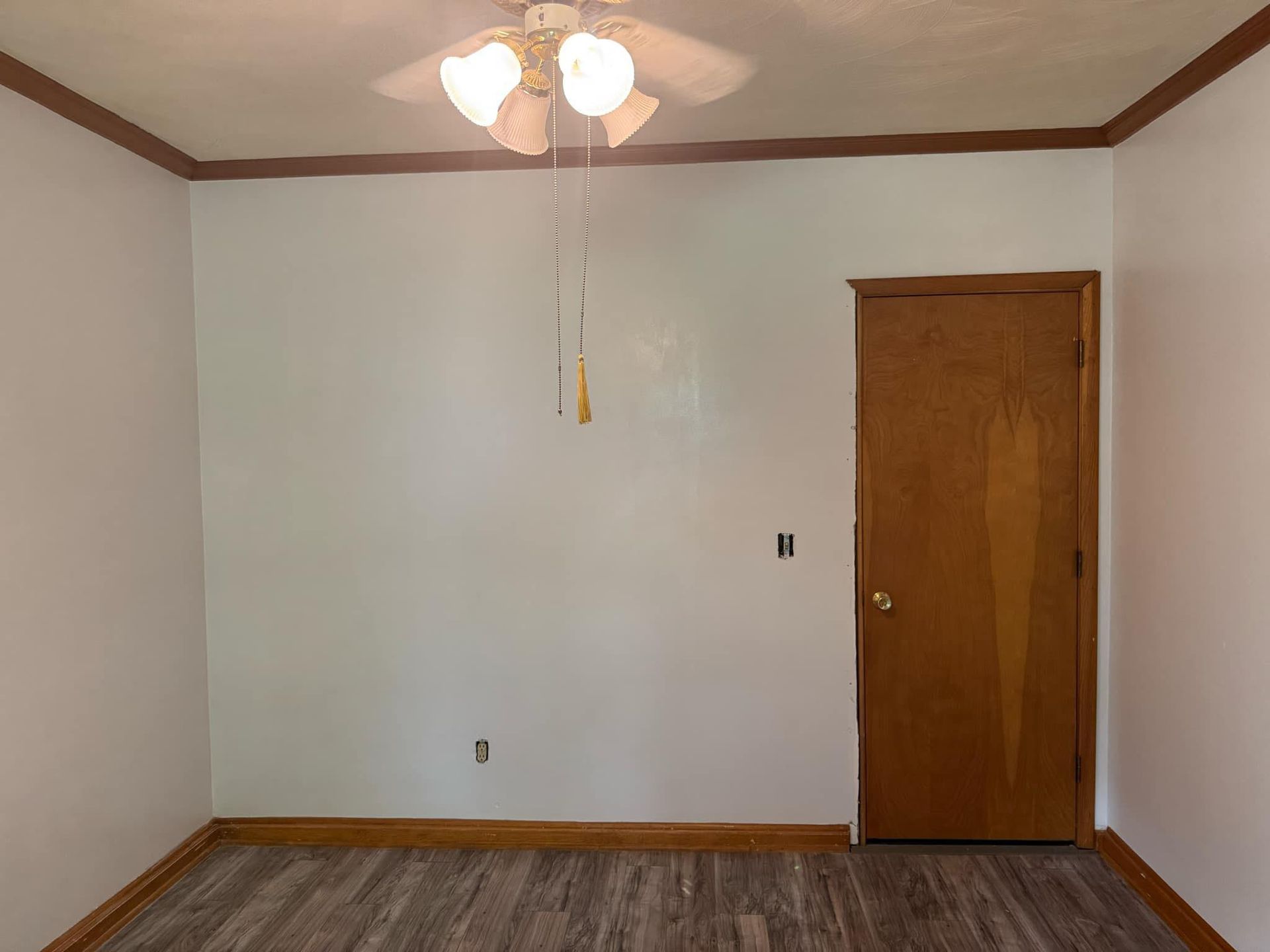 A white-walled room with brown trim, a door, and a ceiling fan; the floor is gray with a wood grain pattern.