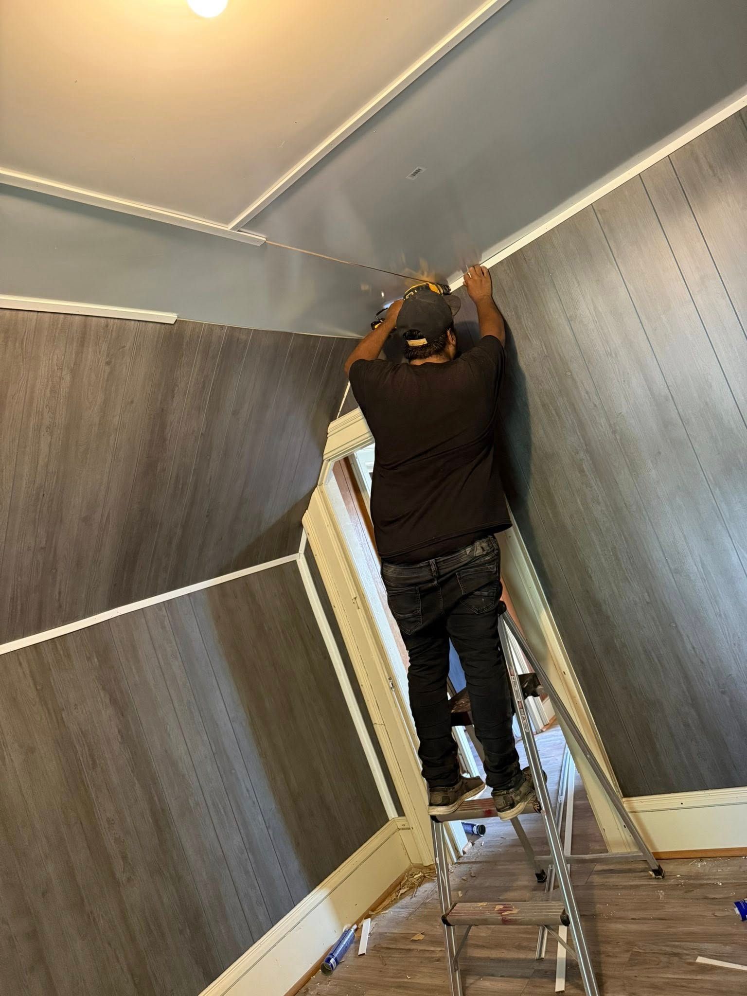 Man on ladder installing gray paneling on a wall, near the ceiling, interior setting.