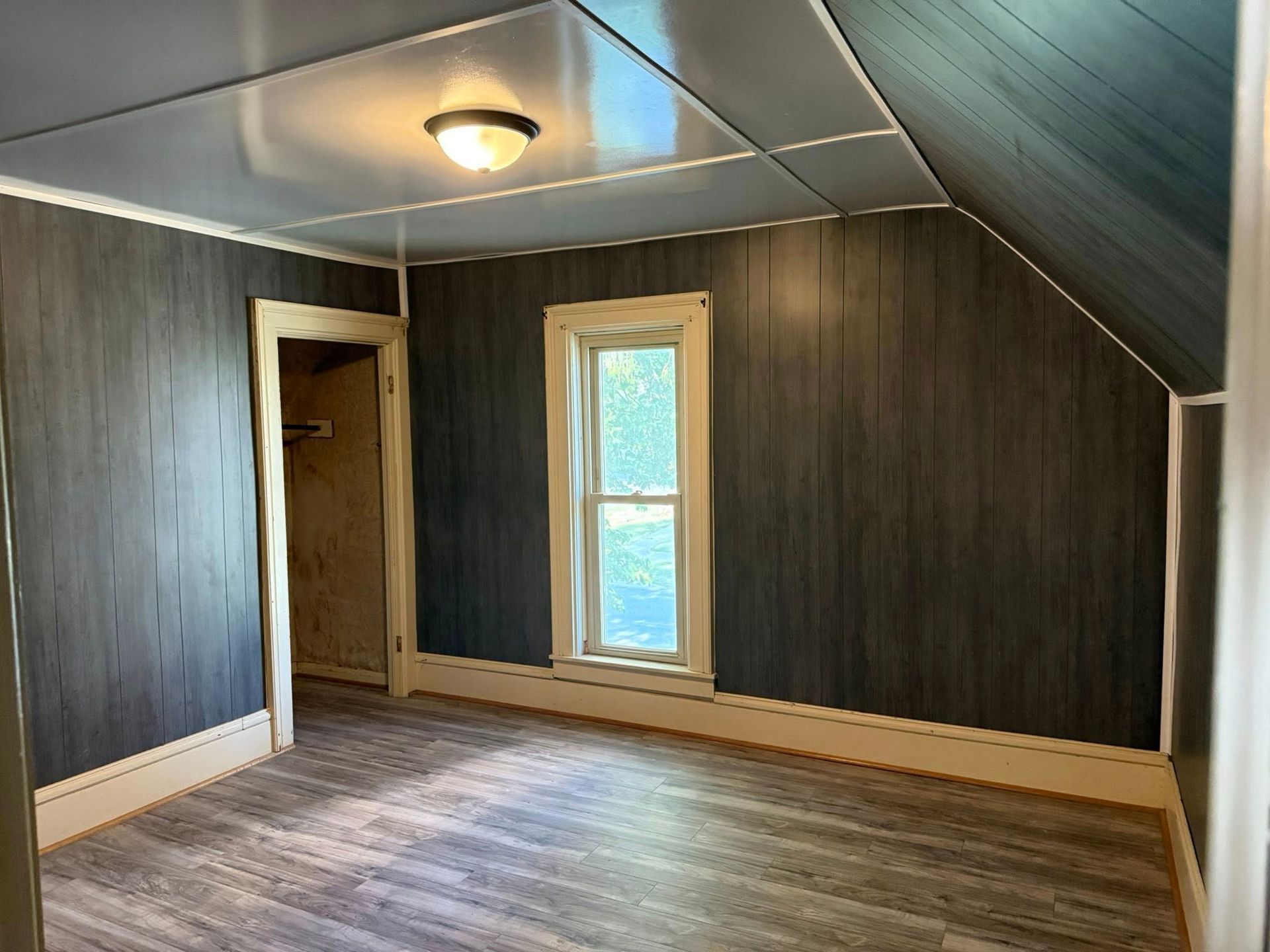 Empty room with gray wood-paneled walls, angled ceiling, a window, and an open doorway.