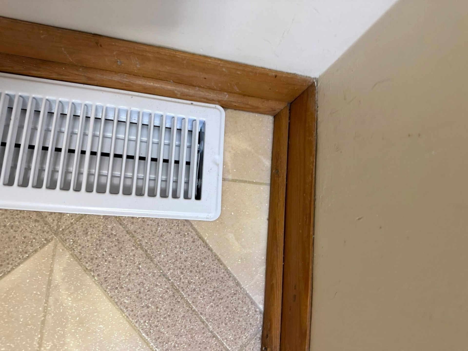 White air vent in floor against wooden trim and tan wall.