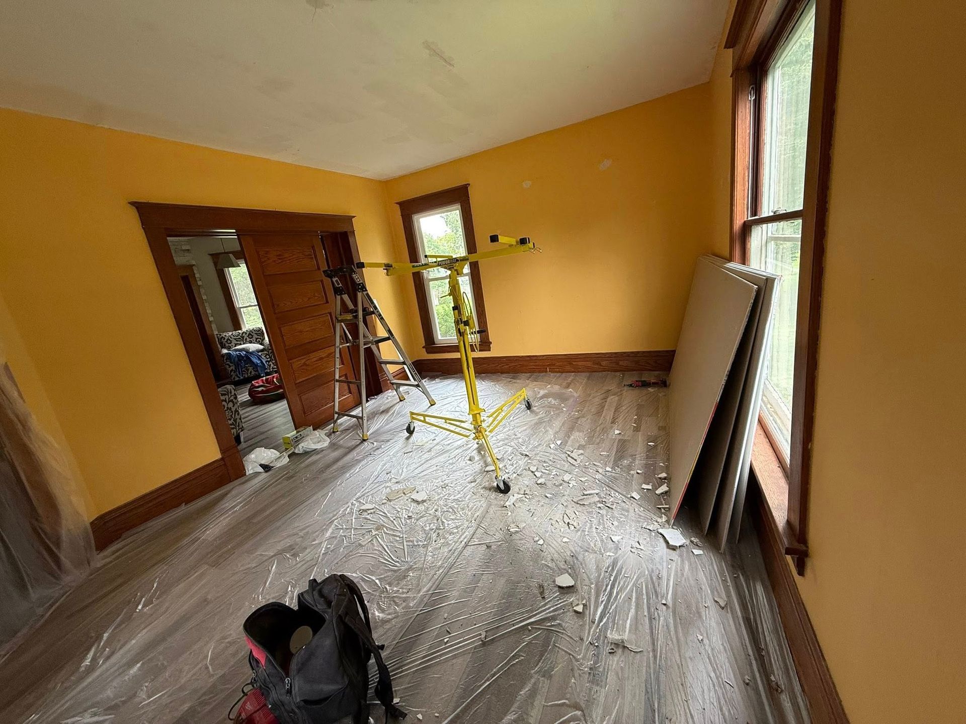 Room interior in renovation with yellow walls, wood trim, plastic floor covering, and a work light.