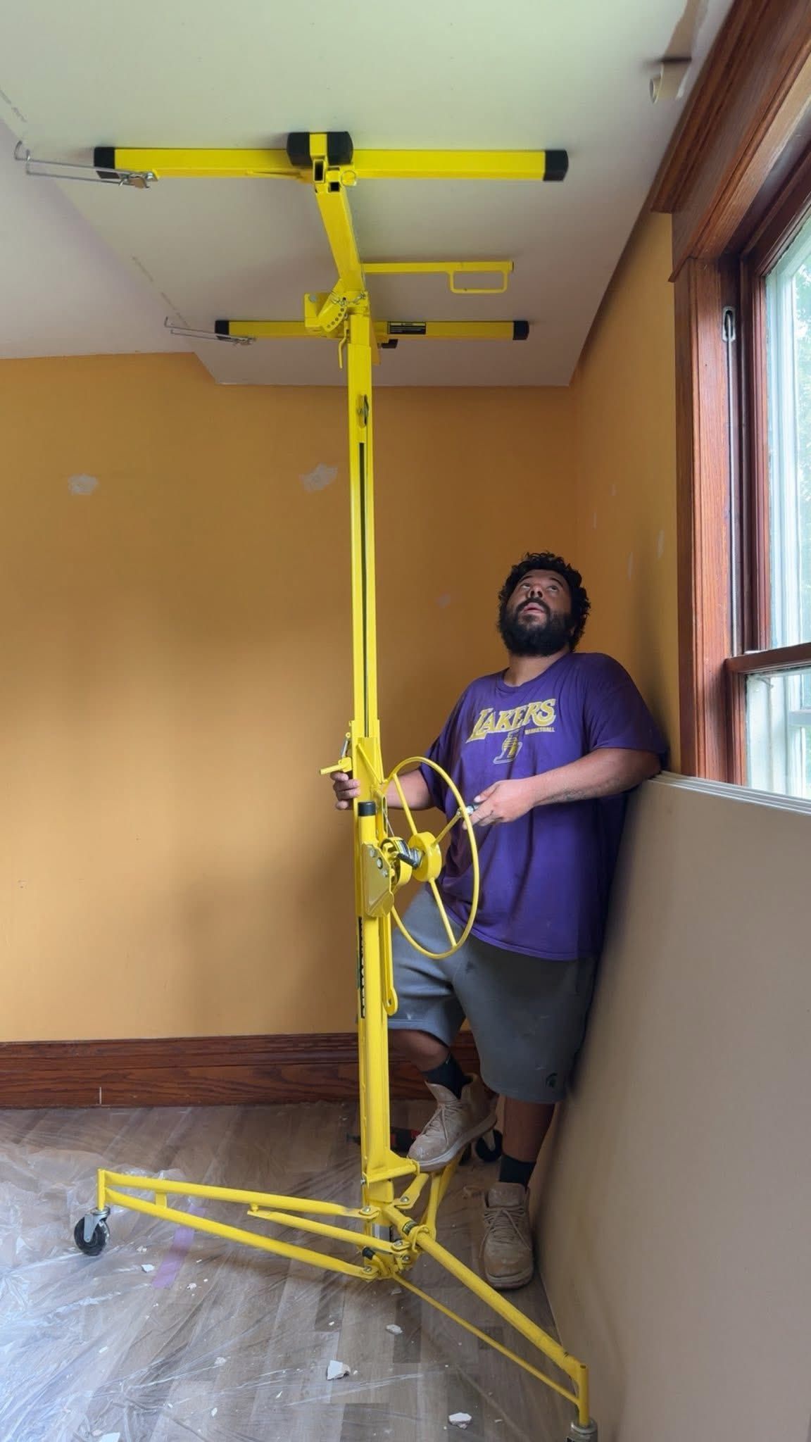 Man uses yellow drywall lift in a room. He leans on the wall near a window, looking up.