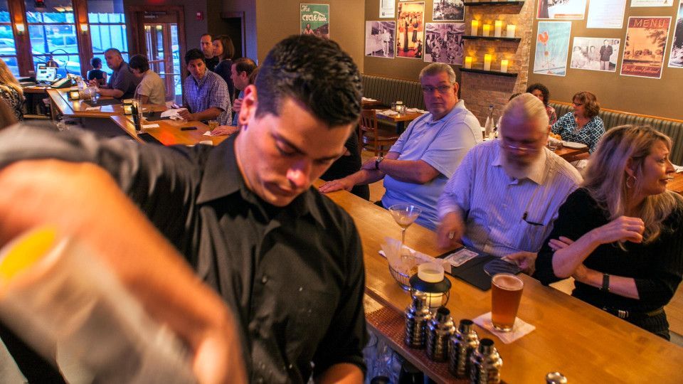 Bartender mixing drinks at a bar, customers seated, soft lighting, and warm tones.