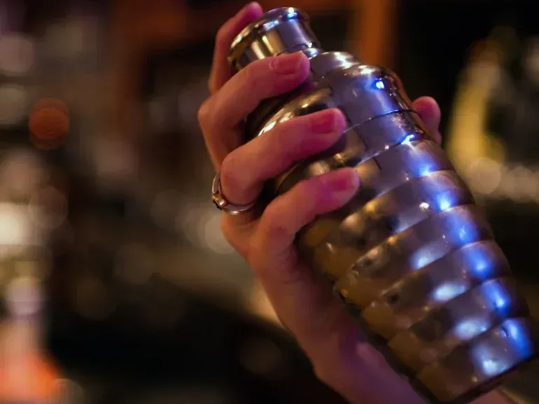 Person shaking a cocktail shaker, blurry bar background.