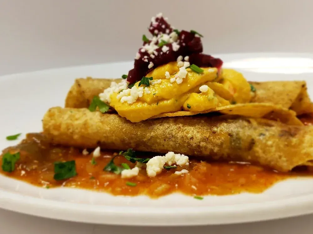 Crispy rolled tacos with salsa, topped with a yellow sauce, beet pieces, and cheese, on a white plate.
