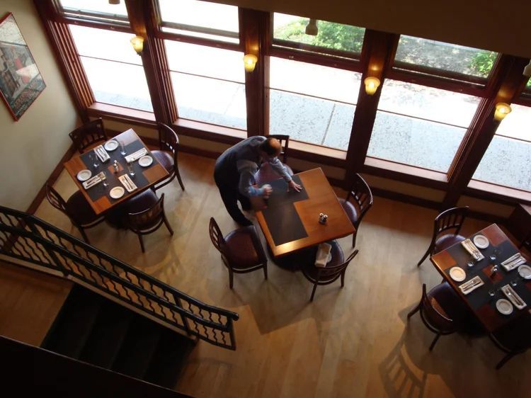 Overhead view of restaurant tables near a large window, one person at a table, another approaching.