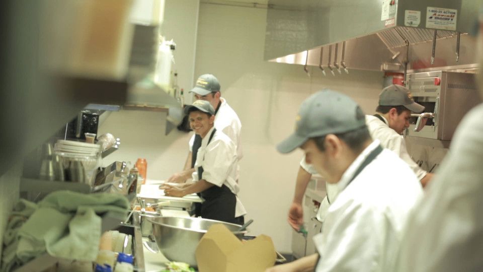Chefs working in a restaurant kitchen. One smiles while cutting food. Stainless steel, bright lighting.