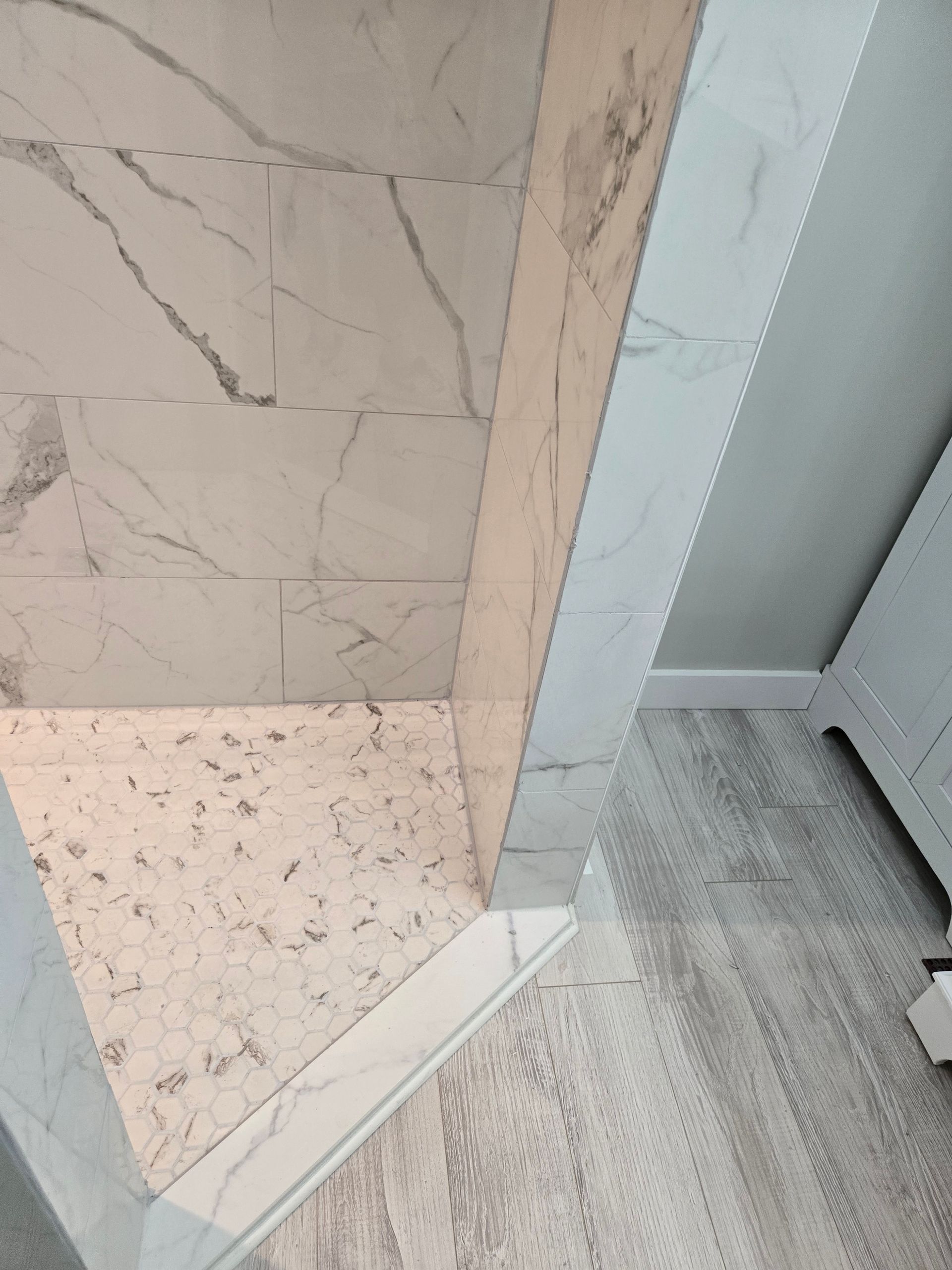 Shower with marble-look tile walls, a stone-like floor, and a white border, adjacent to a light wood-look floor.