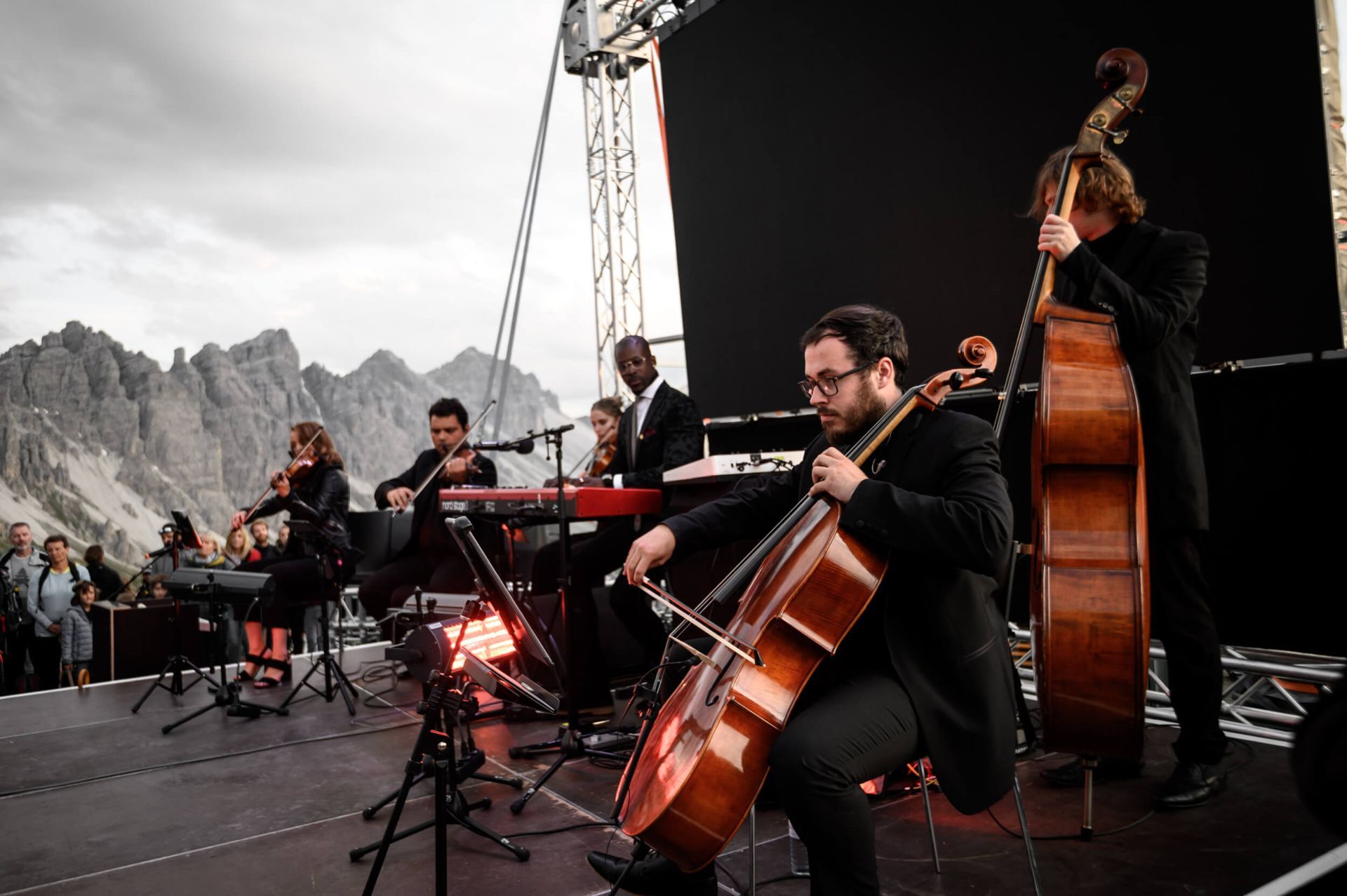 String ensemble performing outdoors with mountains in the background. Musicians playing string instruments, keyboard.