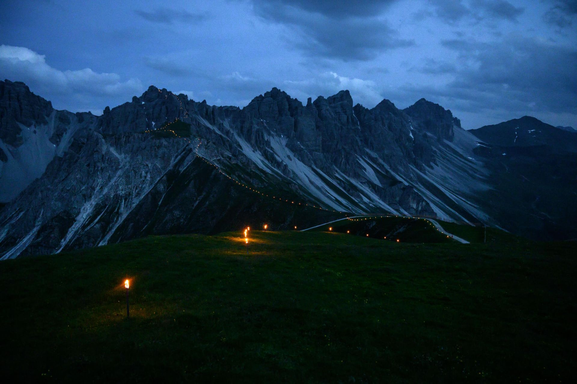 Dark mountain range at dusk, lit by scattered lights on a green hillside under a cloudy sky.