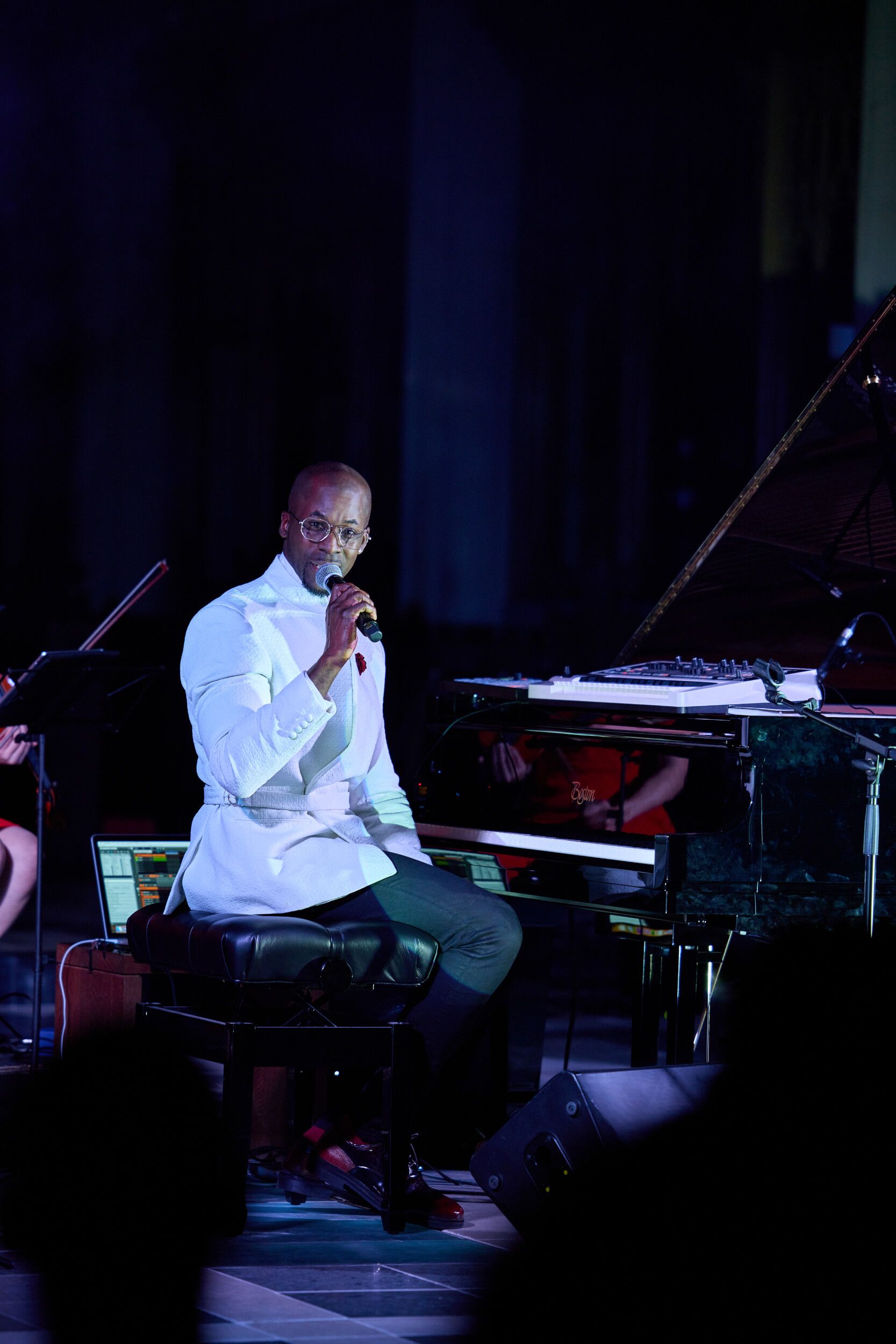 Man singing, seated at a piano on stage, holding a microphone. Dark background with stage lighting.