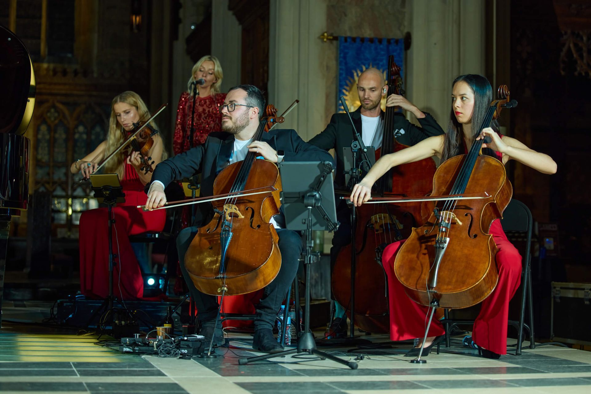 A musical group performing in a dimly lit church. Two cellists, a violinist, singer, and double bassist.