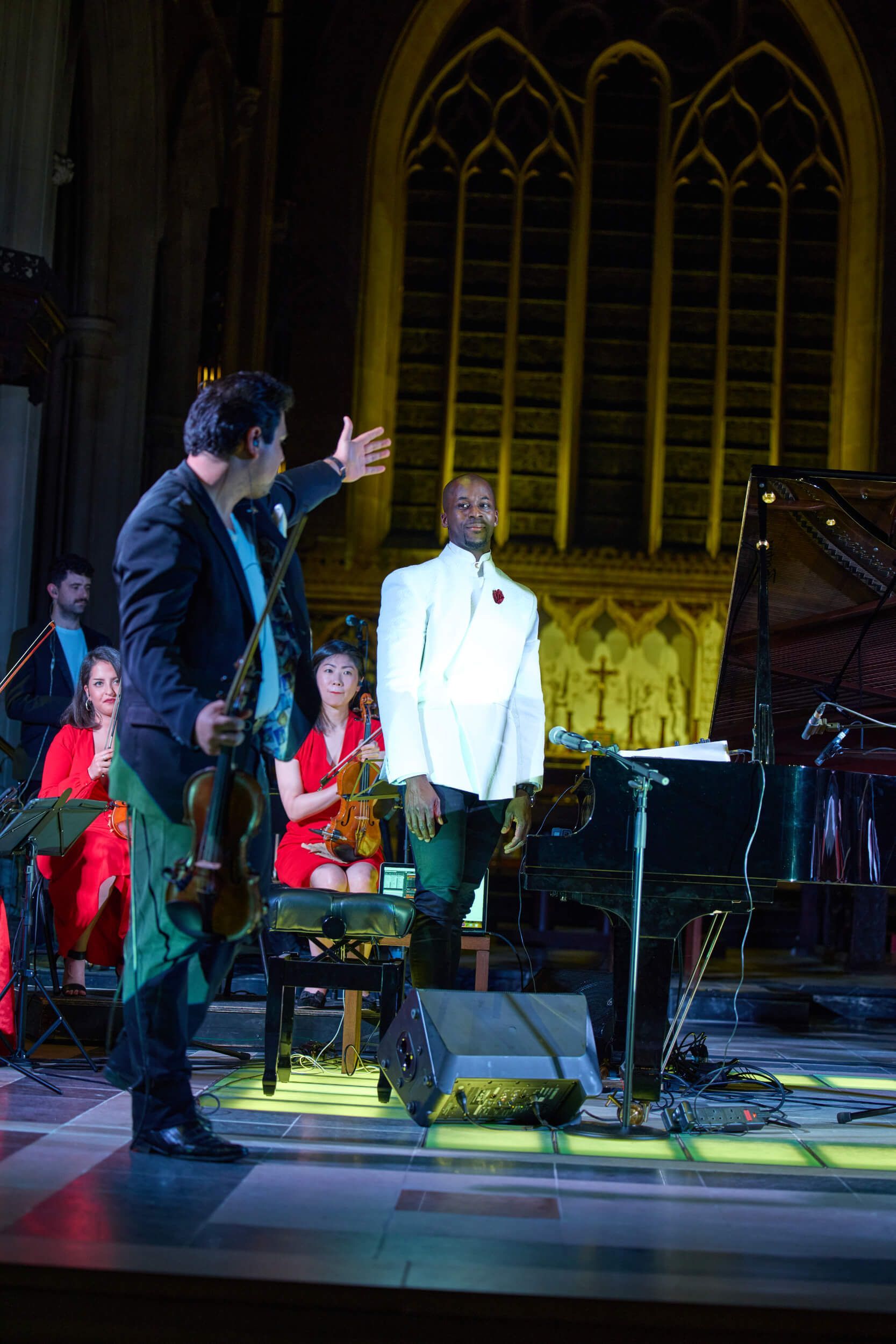 A violinist gestures towards a pianist in white suit on stage. Orchestra in background.
