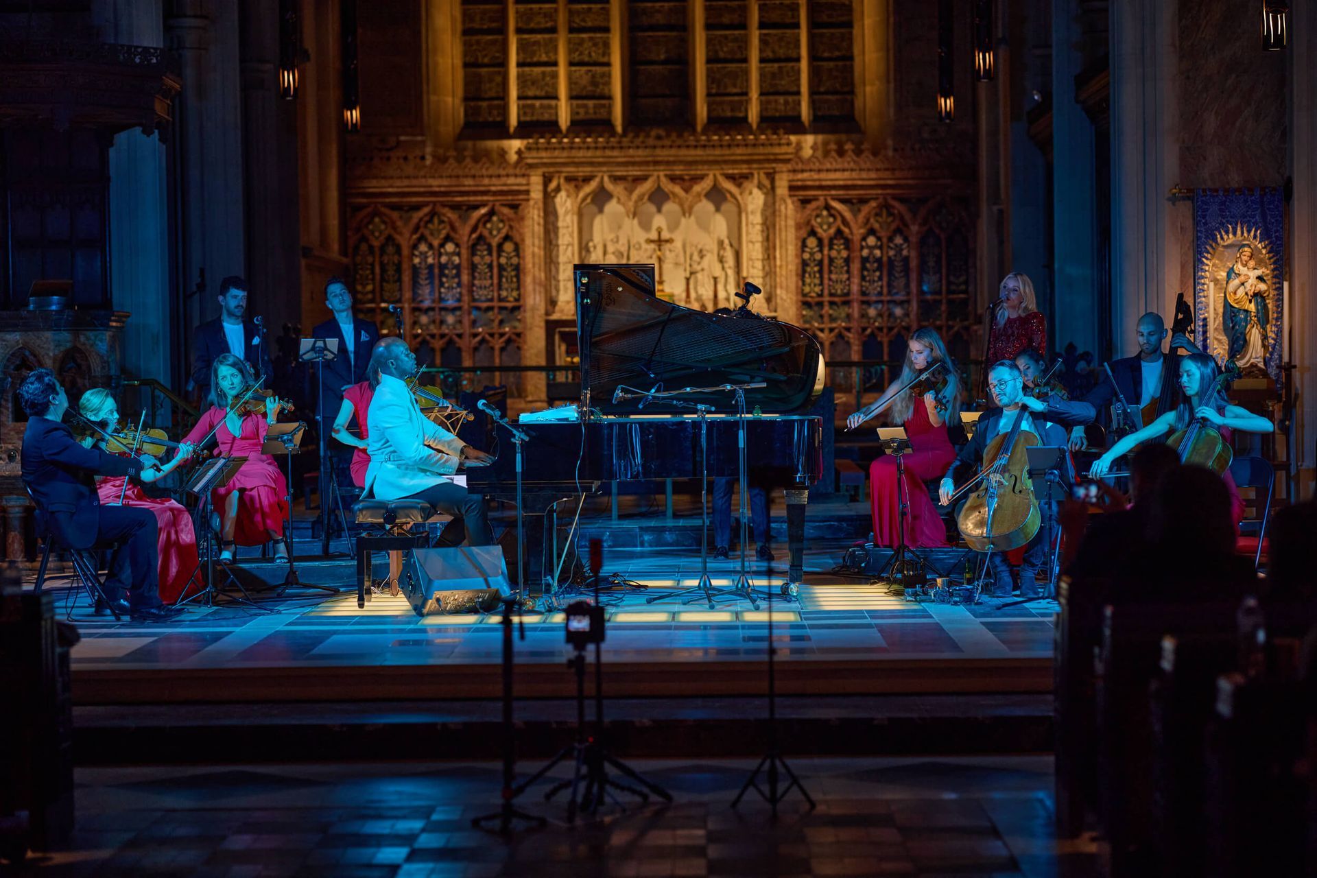 An orchestra performs on a stage in a church. Musicians in formal wear play instruments under stage lights.