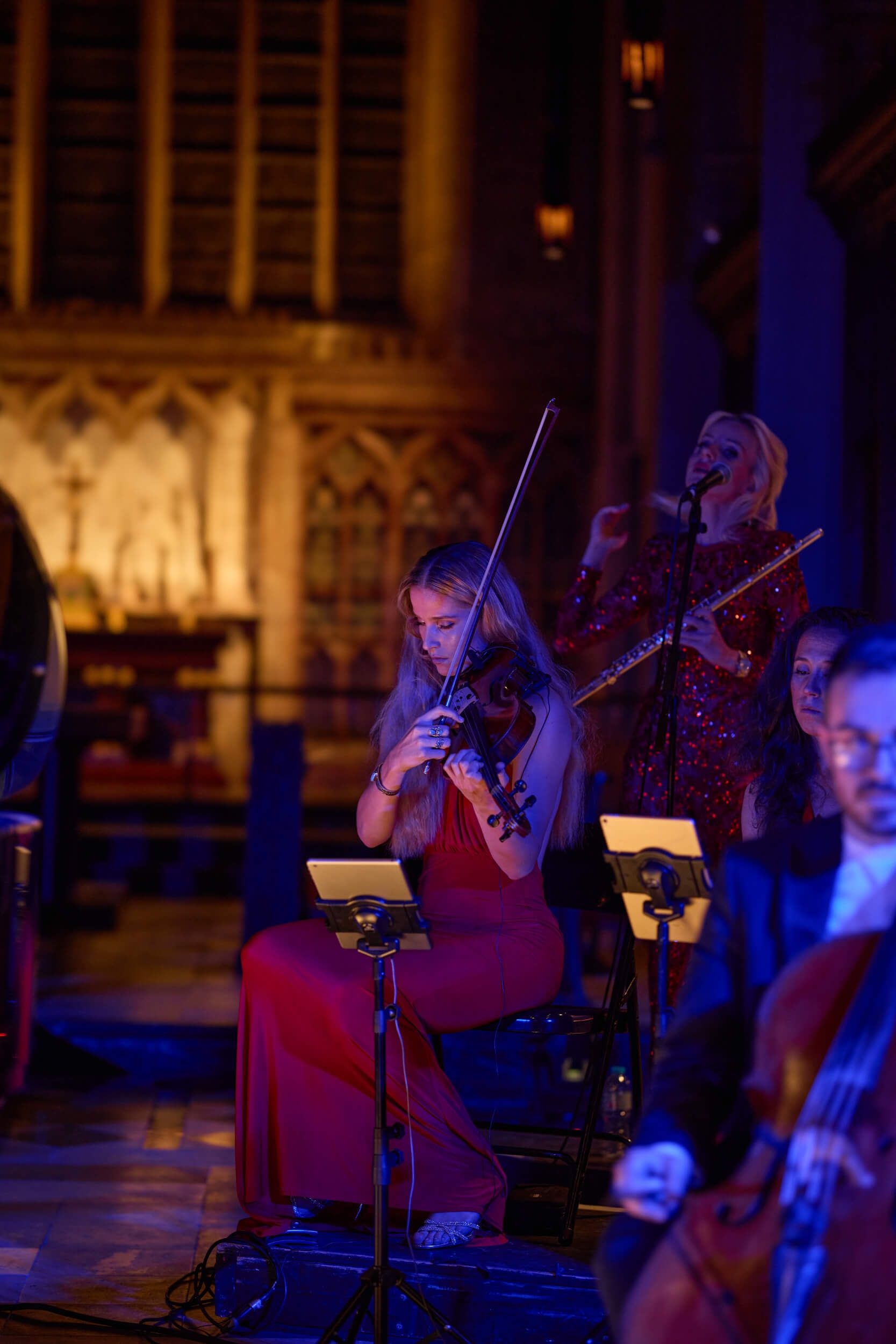 Violinist in red dress plays in an ornate church, with orchestra members and music stands.