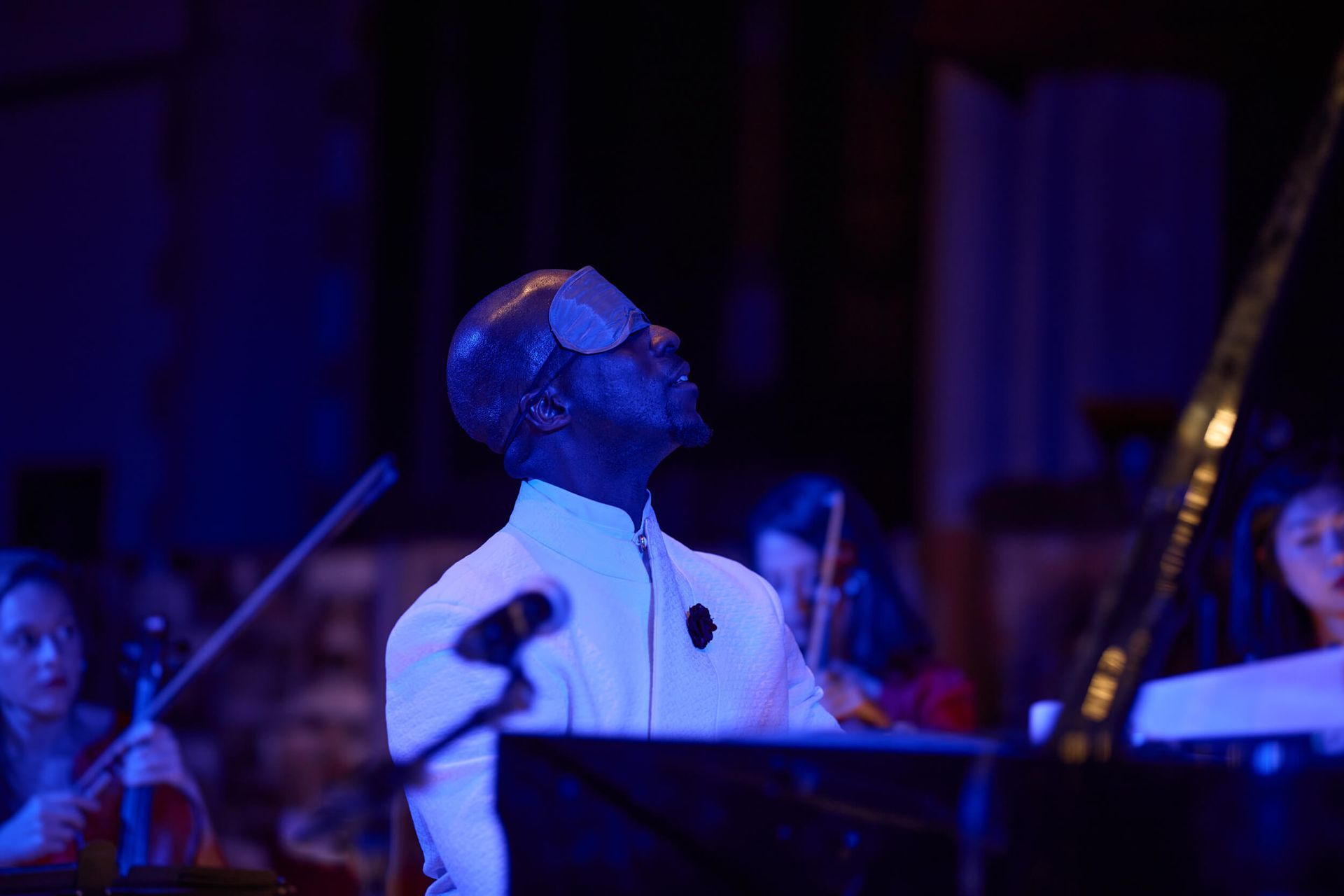 Pianist wearing eye covering, performing with an orchestra on stage, lit with blue light.