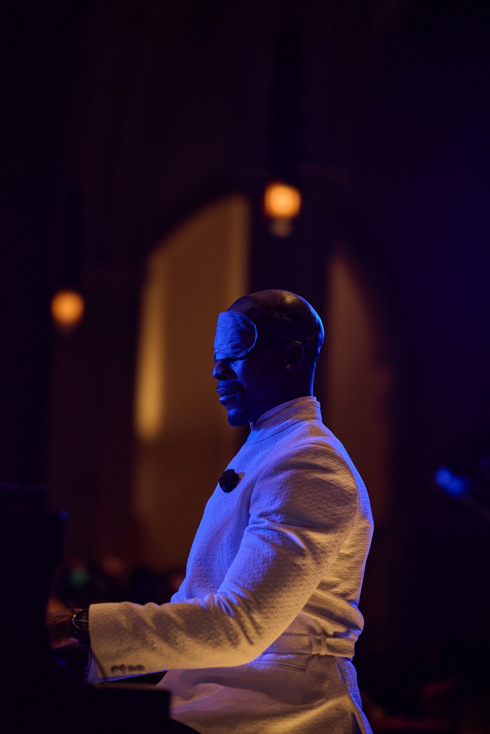Pianist in a white suit illuminated by stage lights. He is seen playing, his head slightly turned.