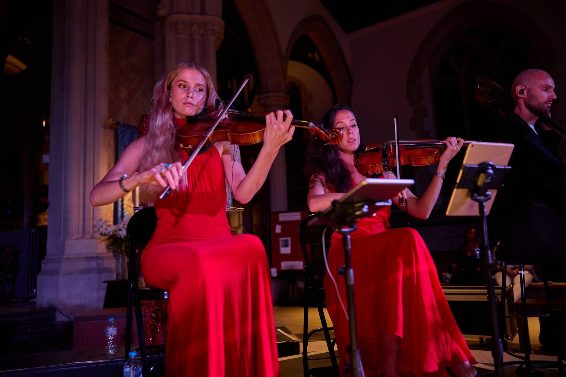 Two violinists in red dresses perform on a stage with a man in the background. Dim lighting.
