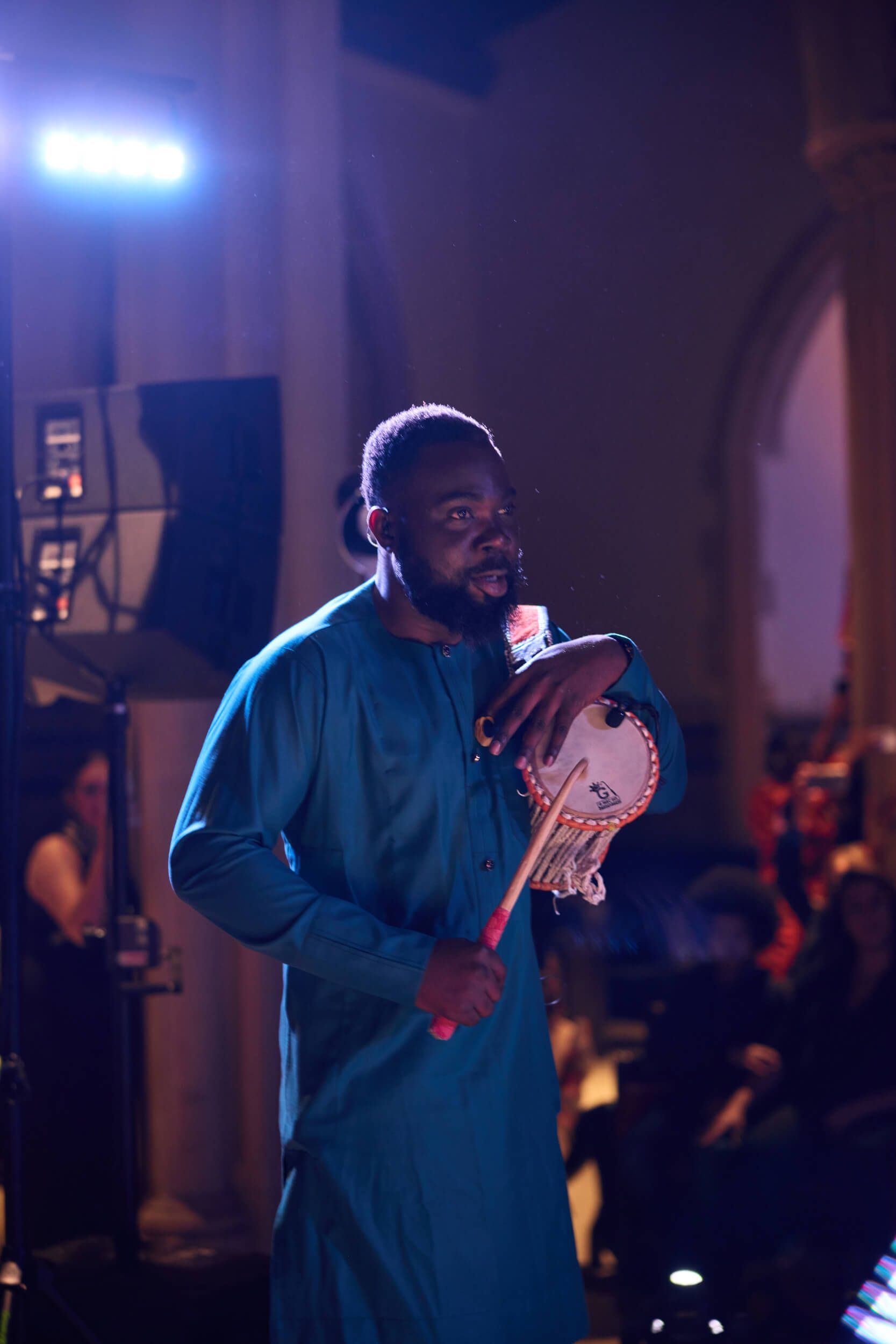 Man in teal shirt plays a stringed instrument onstage, with a blurred audience in the background.