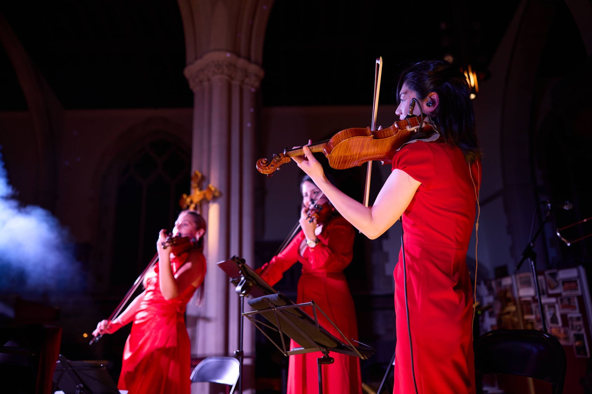 Three violinists in red dresses perform on a stage with a dim, church-like setting.