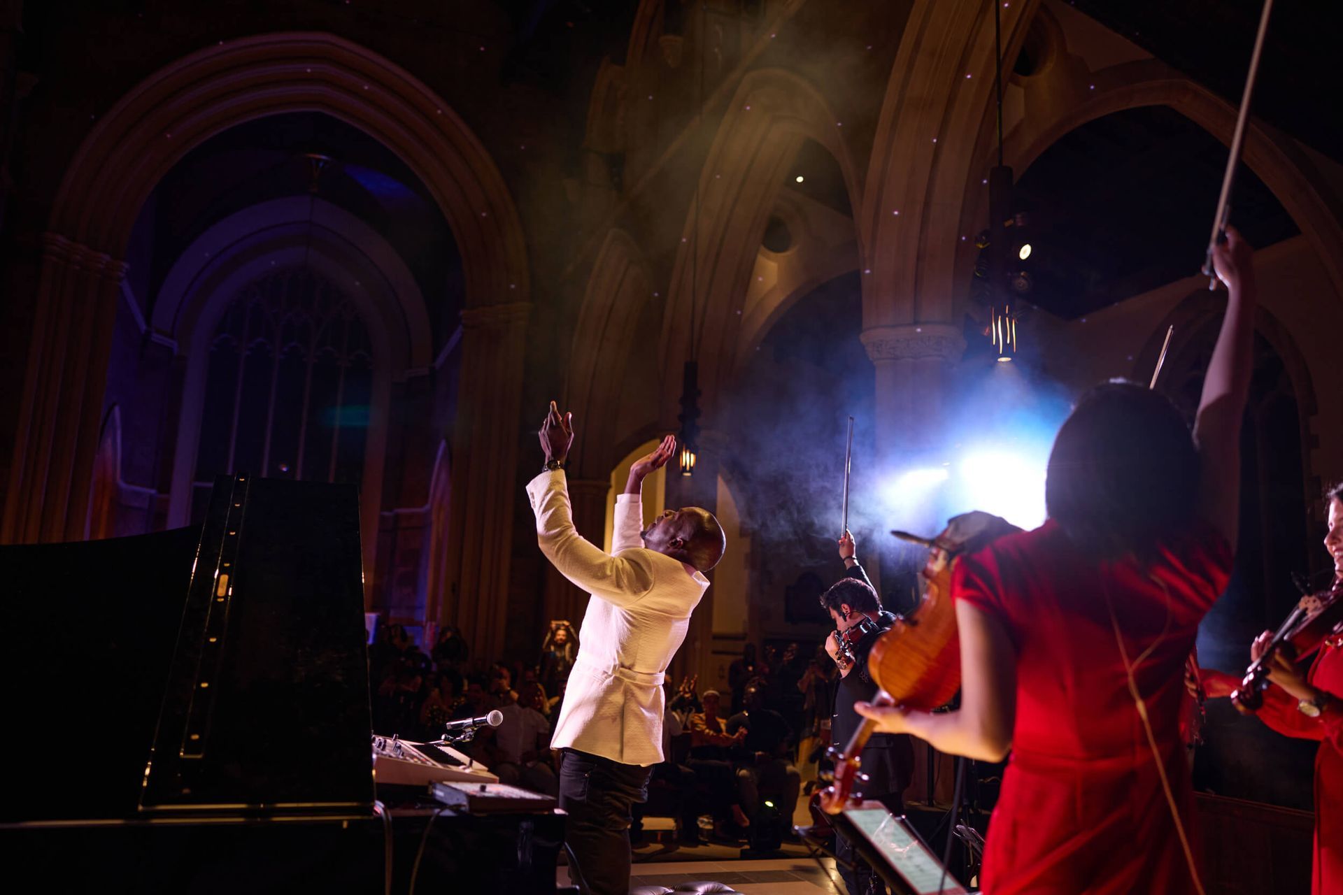 Musicians performing on stage in a gothic church. A man in white raises his hands as a violinist plays.
