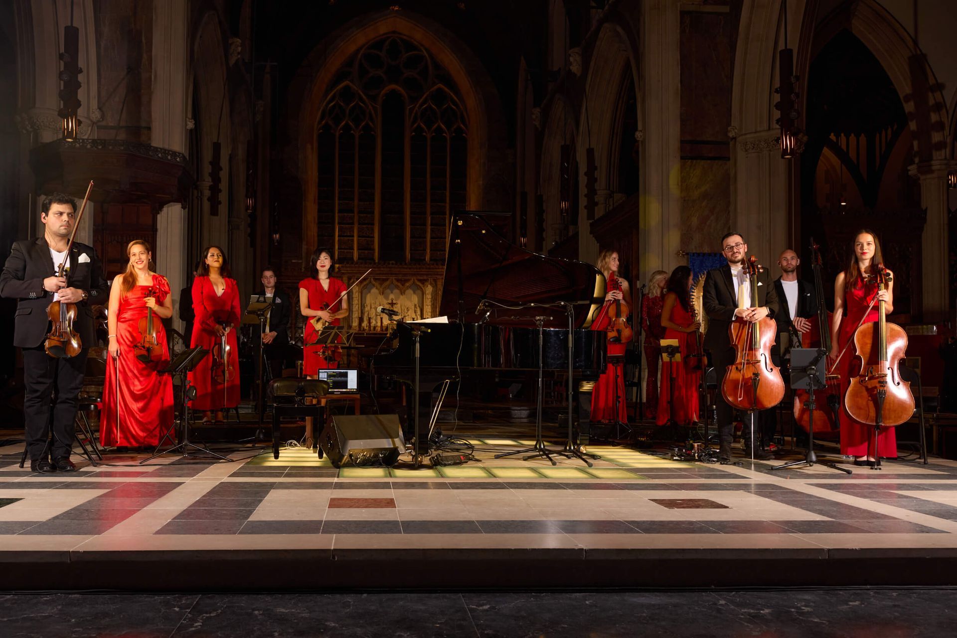 An orchestra performs on stage in a church. Musicians in red and black attire play instruments.