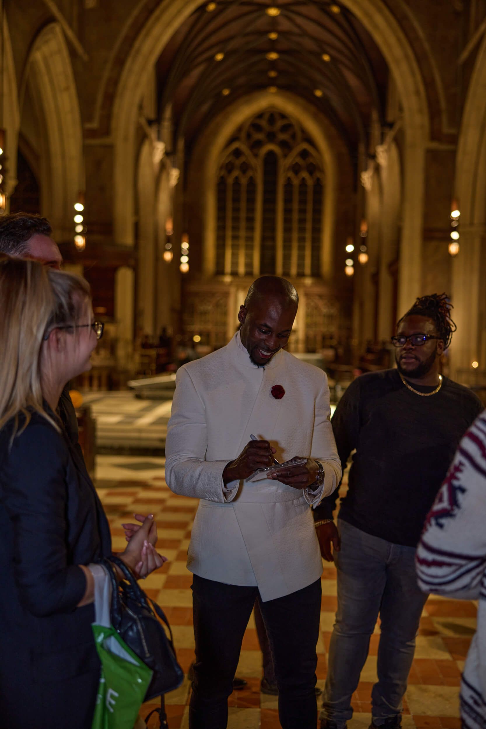 Man in white jacket in a church-like hall, holding object. Others observe, warm lighting, arched window.