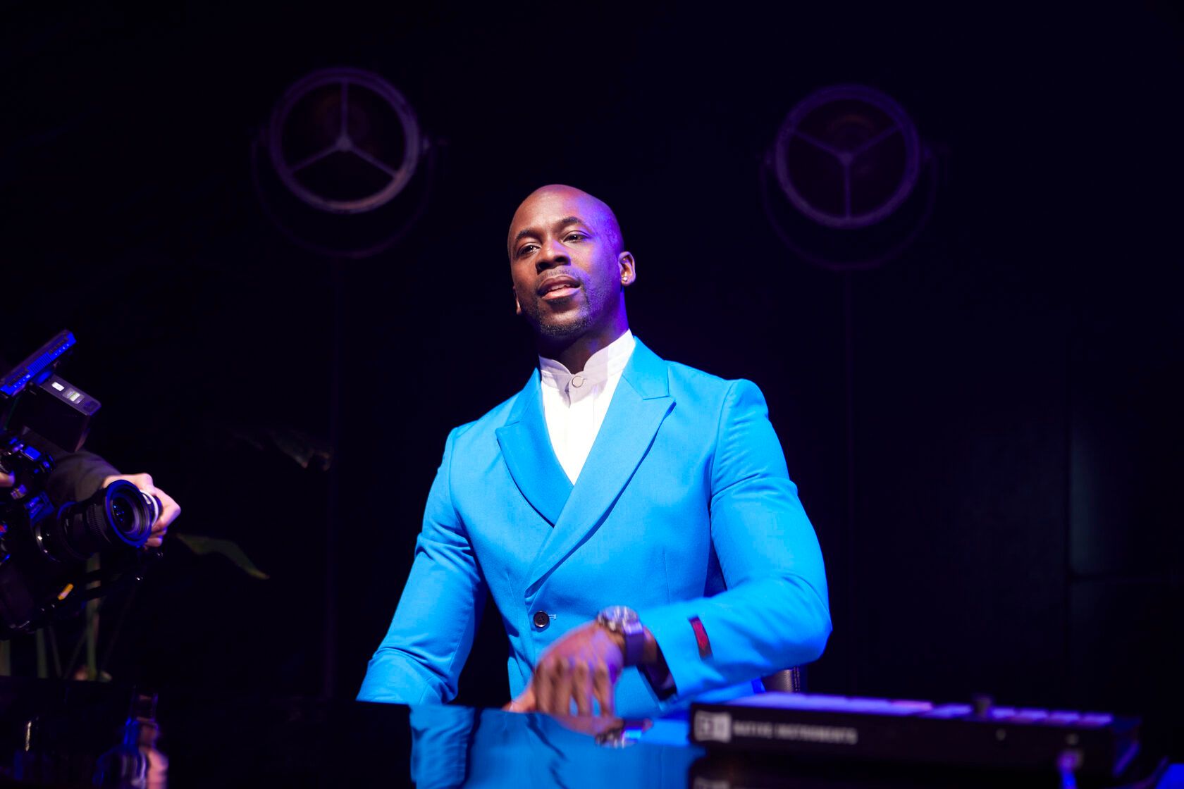 Man in blue suit at a piano, lit stage, looking toward the camera.