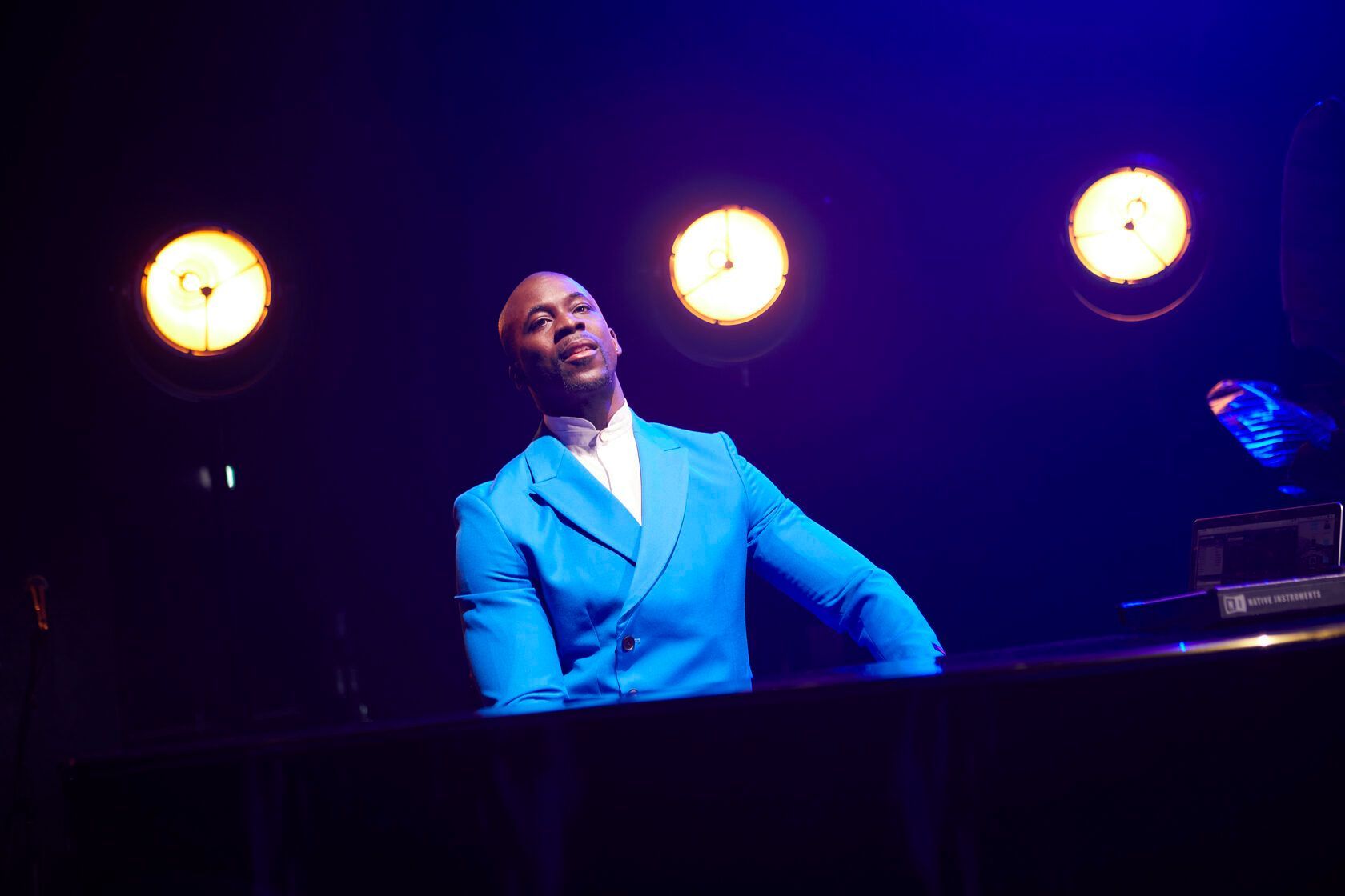 Man in blue suit at a piano, looking upward, lit by stage lights.
