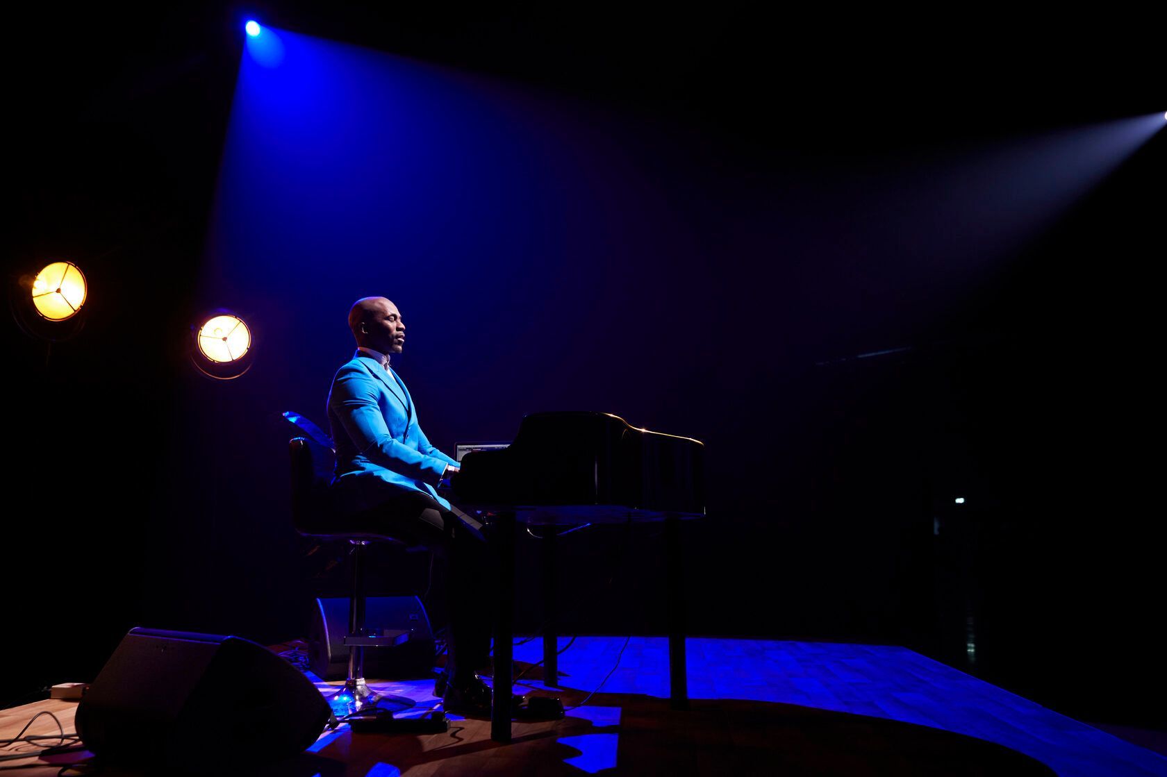 A person in a blue suit plays a grand piano on stage with blue and white spotlights.