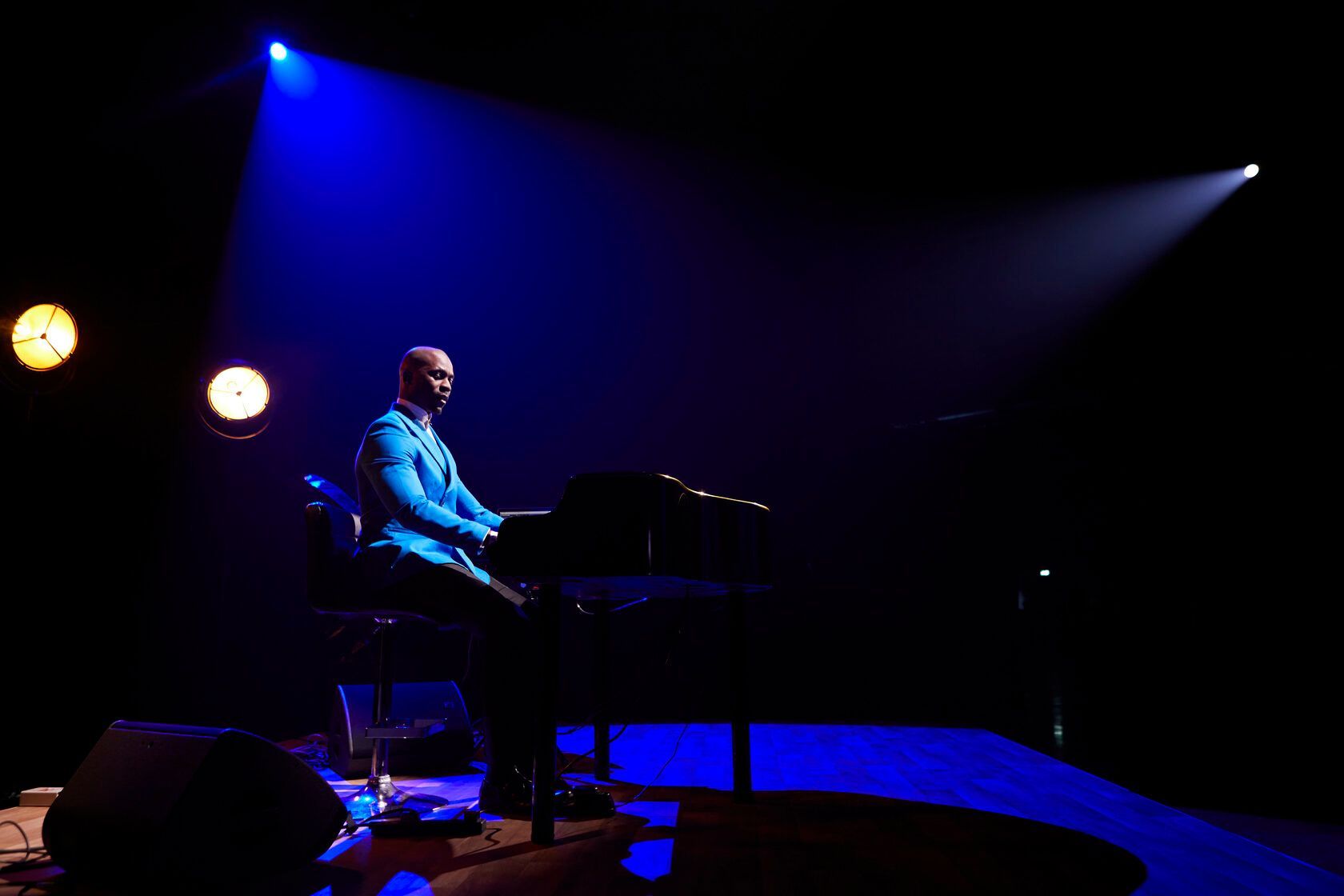 Man in blue suit plays piano on stage, illuminated by blue and white spotlights.