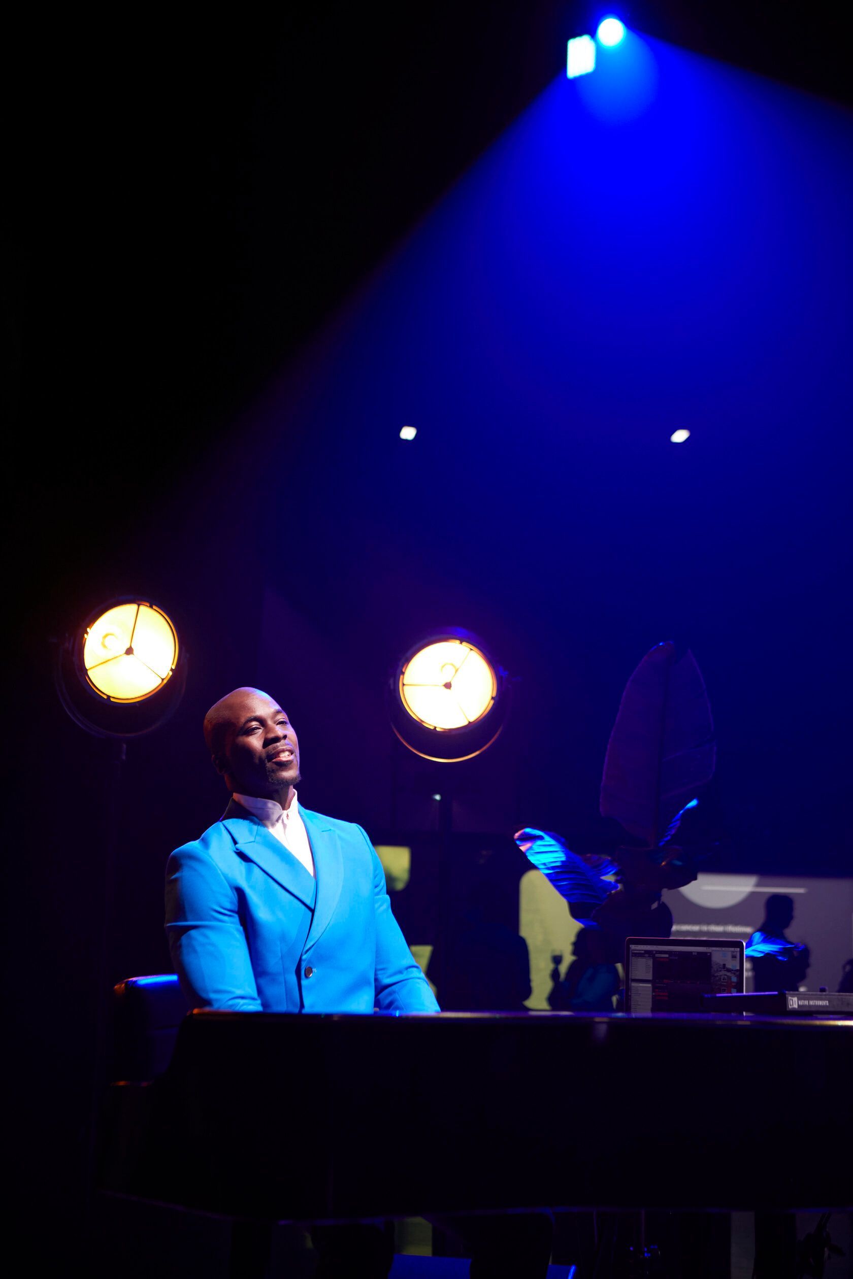 Person in blue suit at a piano under blue and yellow stage lights.