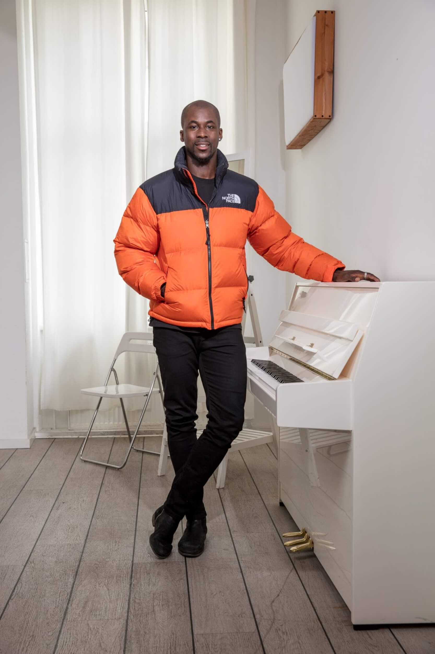 Man in orange puffer jacket stands near a white piano.