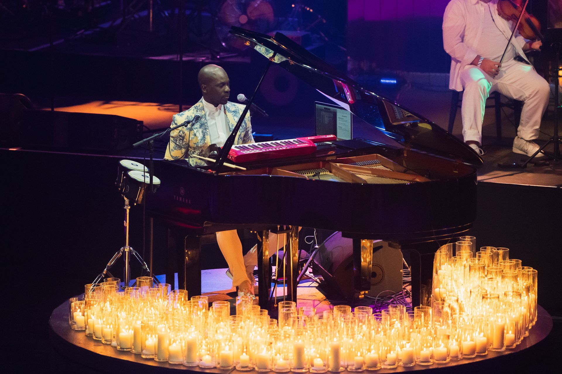 Pianist in colorful shirt performs at grand piano surrounded by lit candles, stage setting.