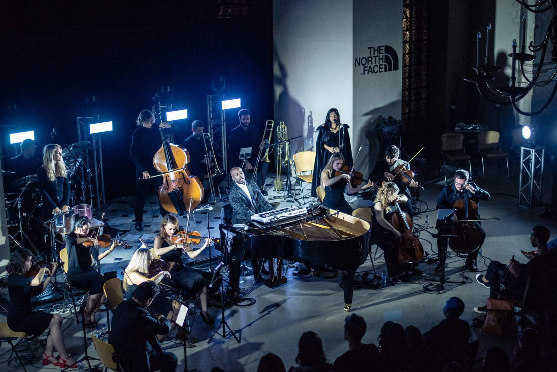 Orchestra performing on stage, lit by spotlights. A pianist, a vocalist, and string instruments are visible