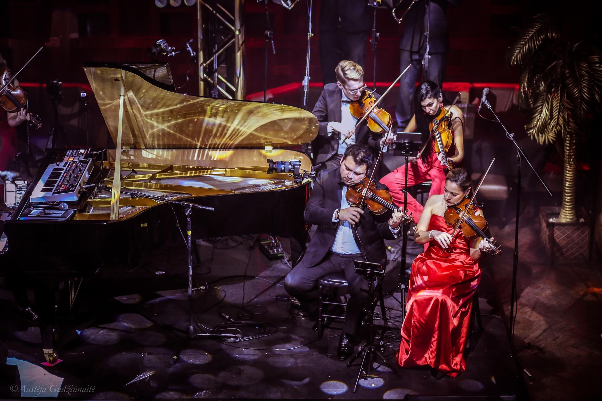 Musical ensemble performing on a stage. A golden piano and violinists in formal attire, stage lit.