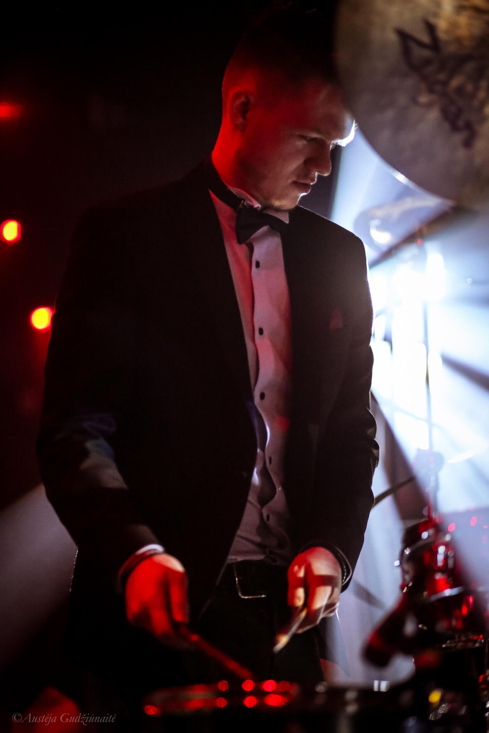 Drummer in a suit performing on stage, lit with red and white lights.