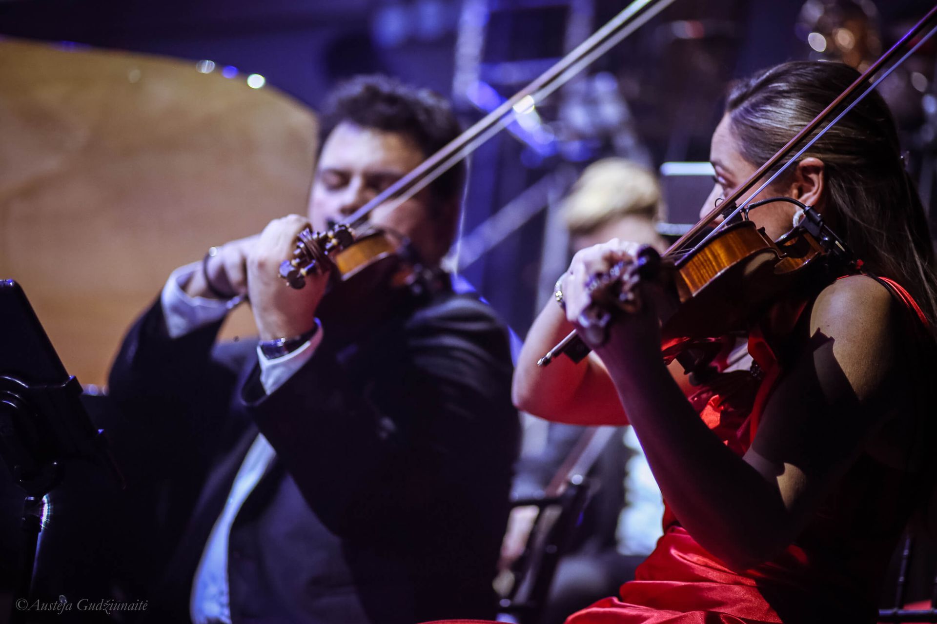 Two violinists in an orchestra, playing. One in a red dress, one in a suit. Stage with blue lighting.