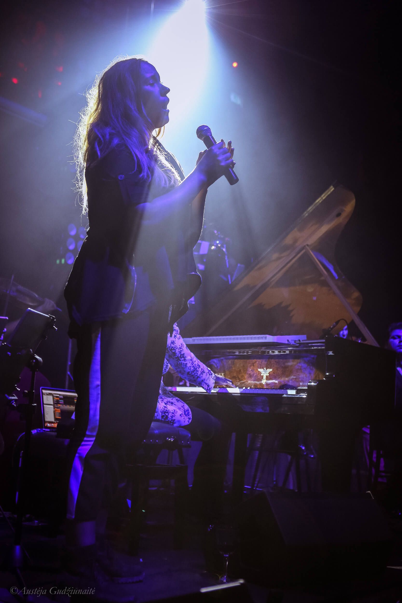 Singer on stage, backlit by a spotlight, holding a microphone. Keyboard and band members are in the background.