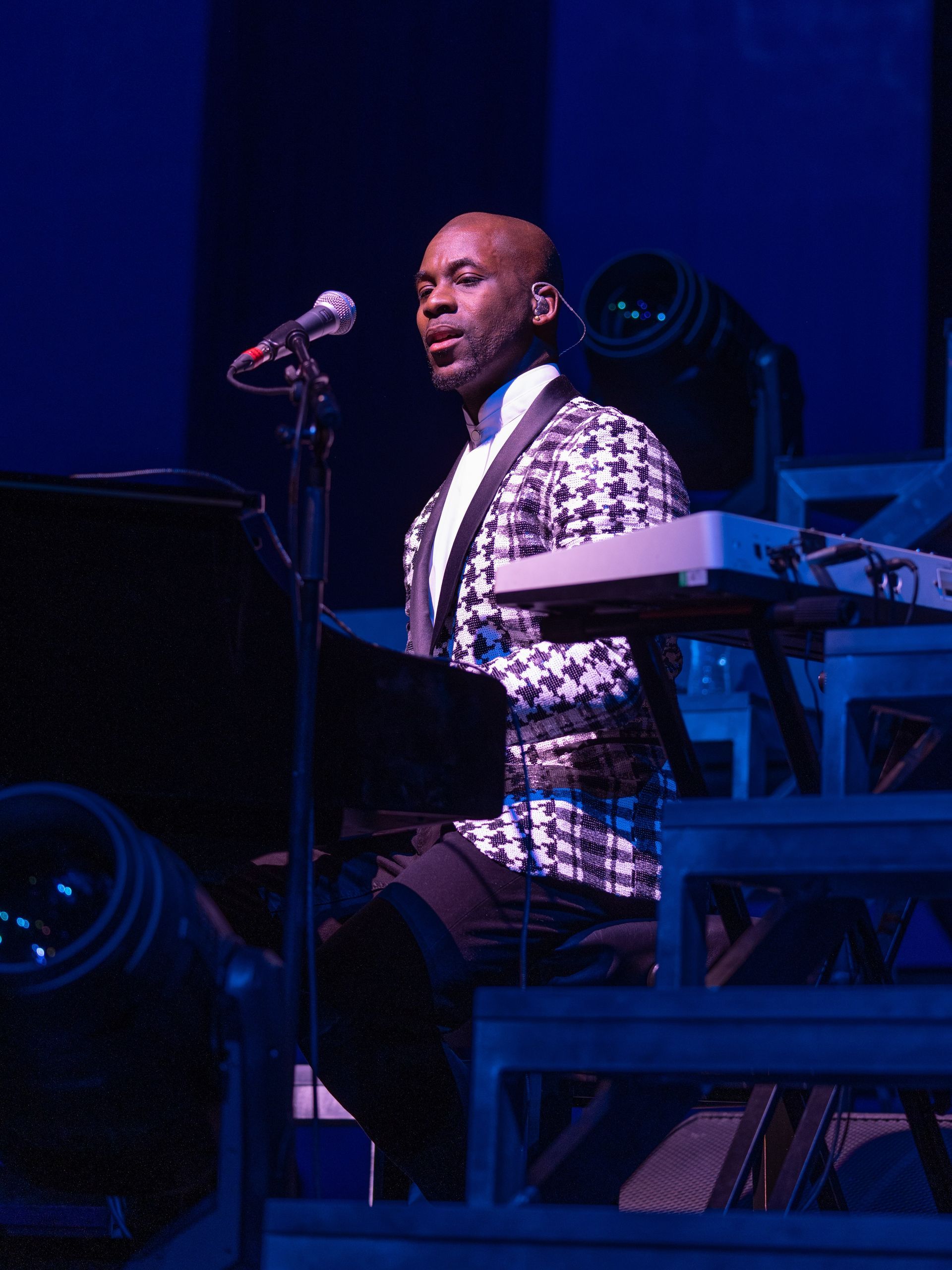 Man singing at a piano, wearing a patterned jacket, on stage with blue lighting.