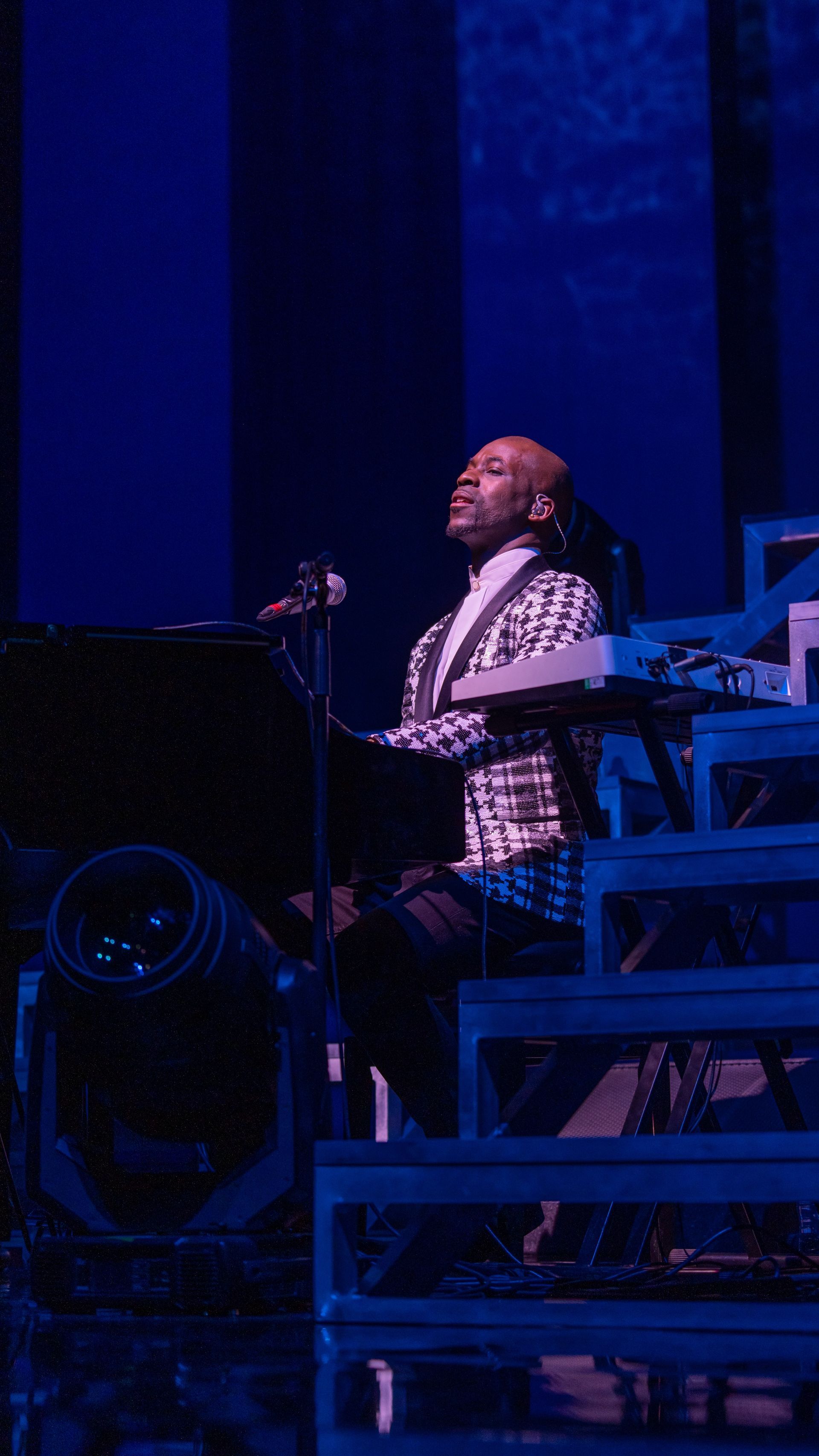 Man in patterned jacket plays keyboard on stage, lit with blue light.